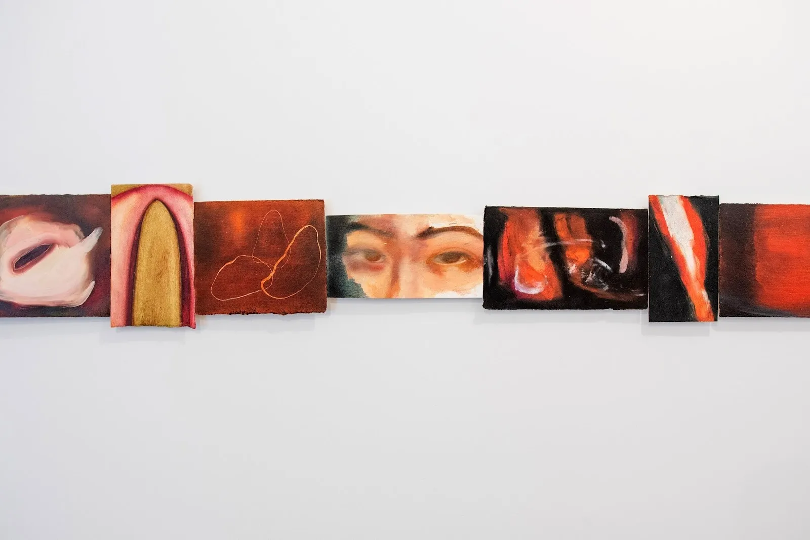 Multiple small paintings of different sizes are hung flush together in a chain on a white wall. Each painting, featuring brown, flesh, red, and black hues, are abstractions or figurative representations.