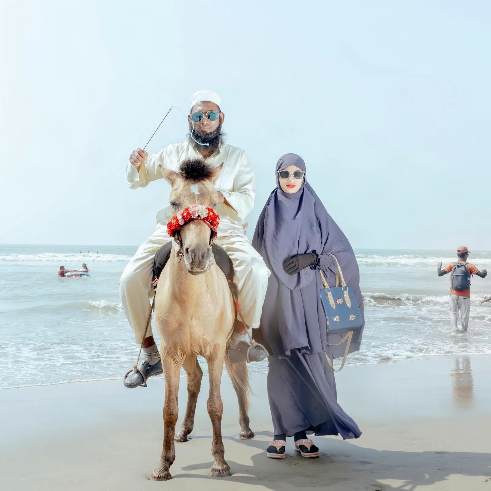 The photograph depicts a man in white riding a horse, posing next to a woman in a monochrome dress and khimar, sunglasses, gloves, a purse, and red lipstick.