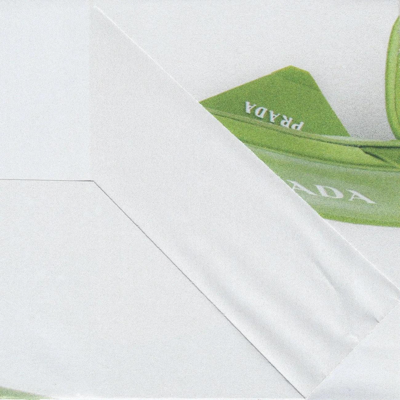 A close-up of a folded white paper surface revealing part of a green Prada bag with the brand name visible.