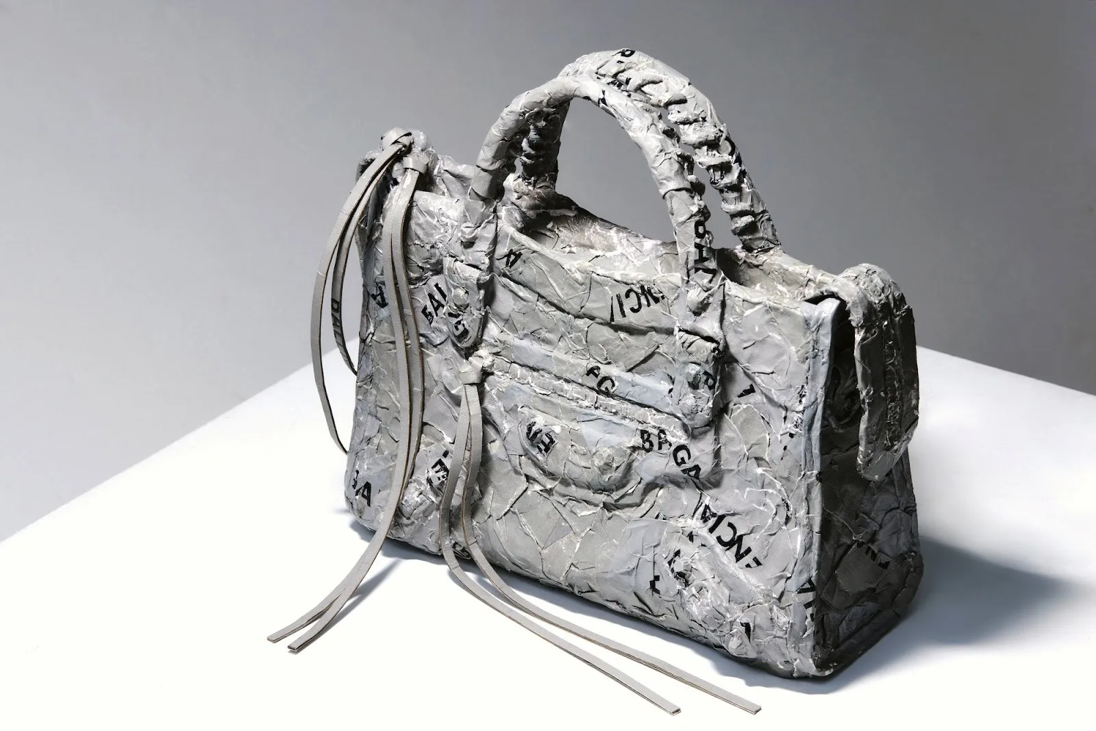 A crumpled-looking gray sculptural handbag with imprinted Balenciaga logos, resembling wrinkled paper or foil.