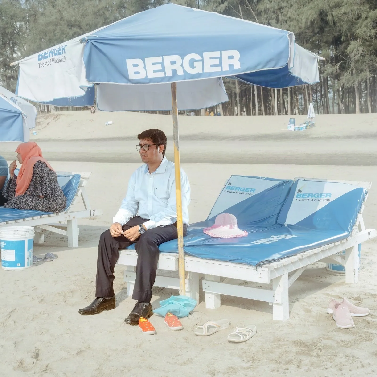 A man in a blue collared shirt, slacks, and dress shoes sits on beach lounge chairs under a blue stand umbrella that reads "BERGER." Around him are children's shoes, sandals, and hats.