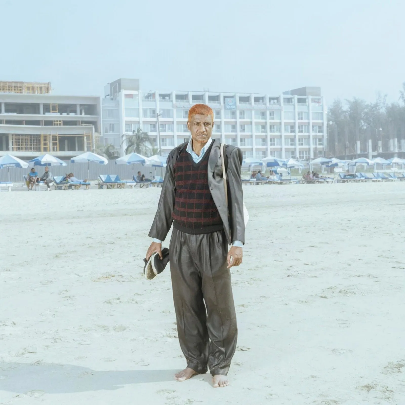 A man wears a silvery suit with a sweater and collared shirt underneath. His hair is cropped and bright orange, and he carries a tote bag and shoes in his hand. He stands barefoot, looking straight at the camera.