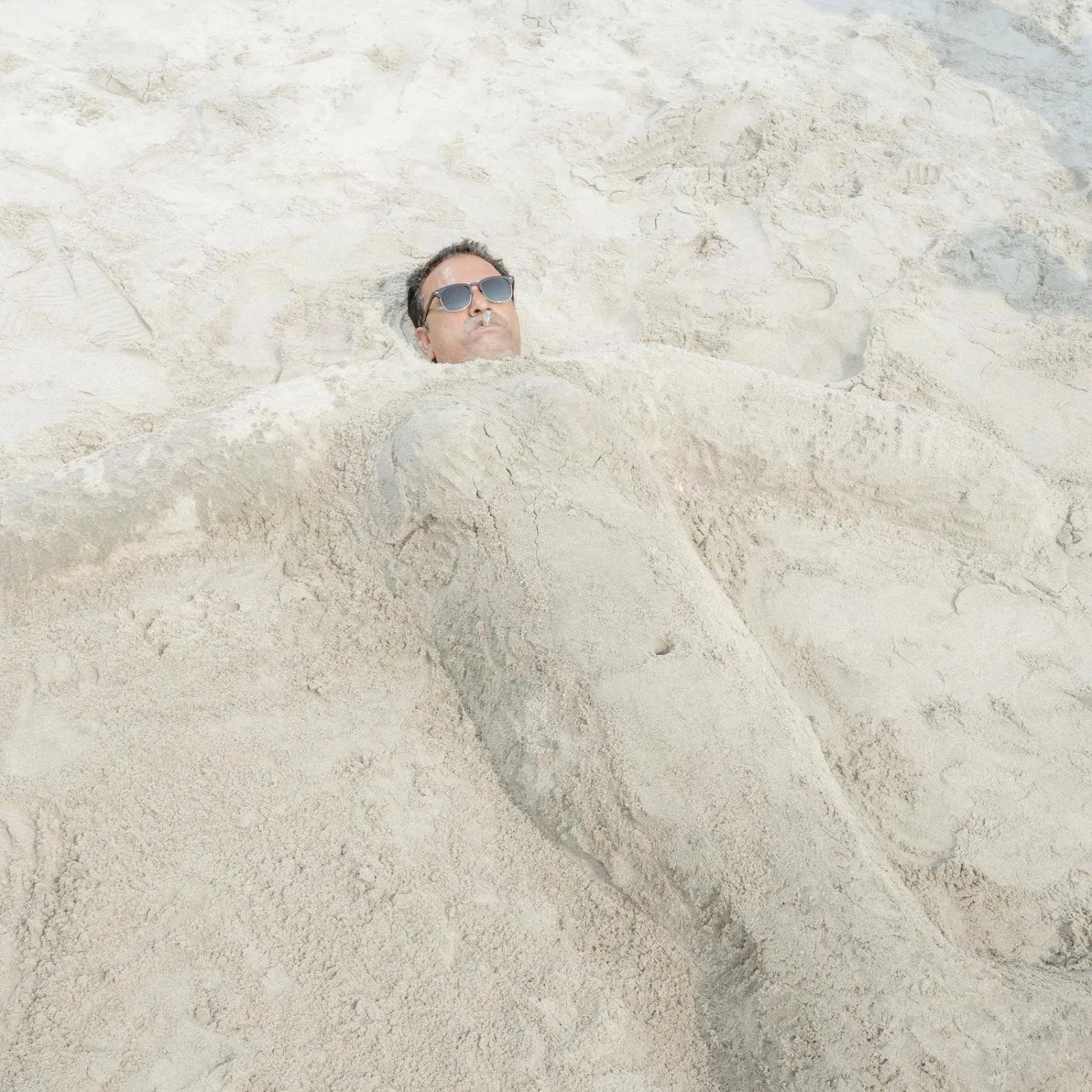 A man, covered in sand to make the outline of a body, wears sunglasses and smokes a cigarette. His entire body is covered in sand, save his face.