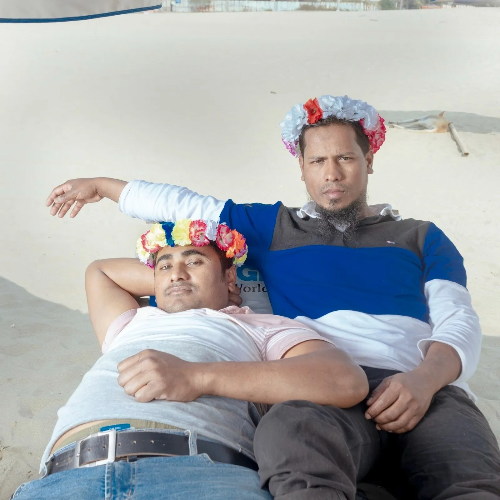 Two men recline on a beach lounge chair. They wear colorful flower crowns and drape over each other comfortably. The look directly at the camera.