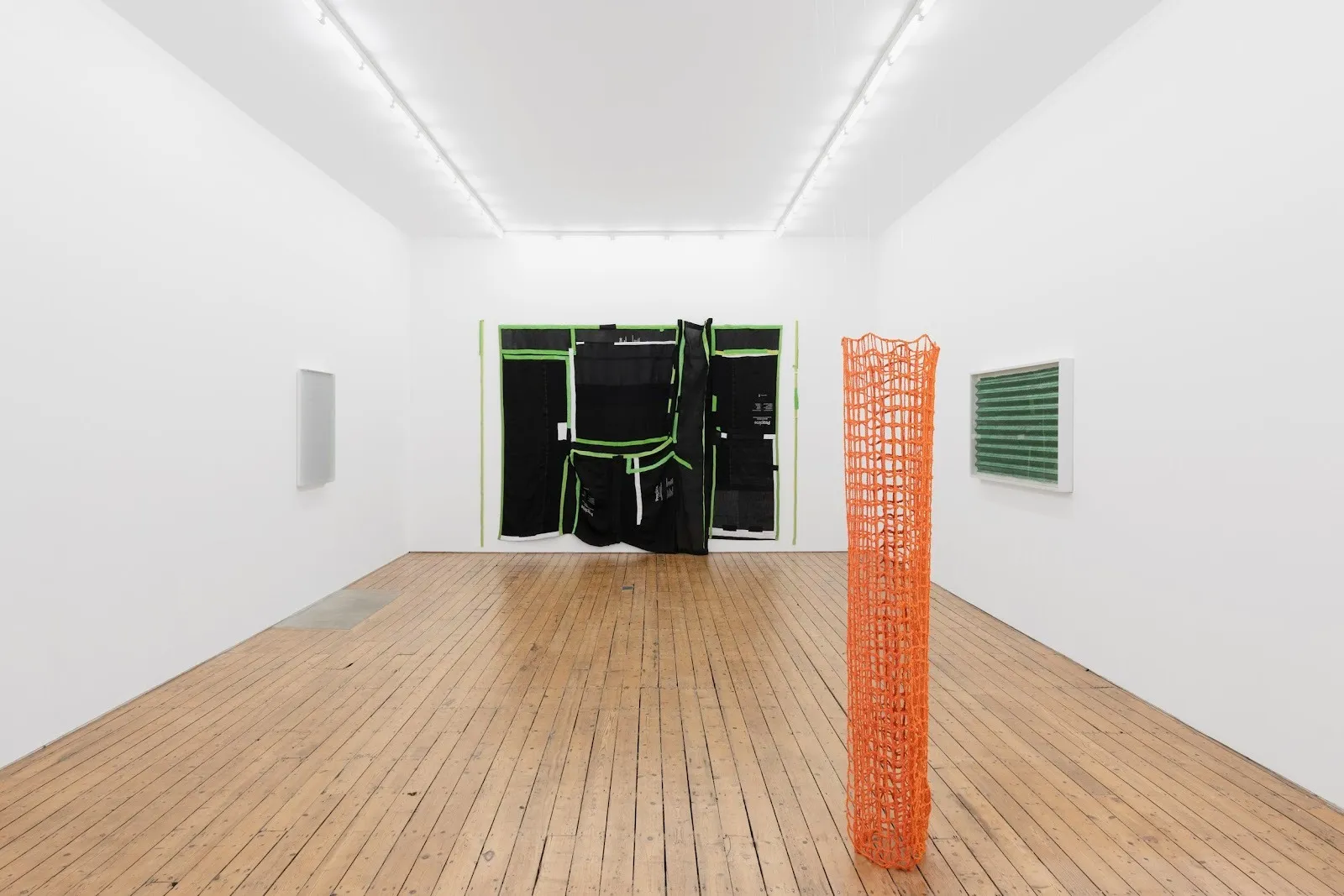 A white gallery room features three wall pieces and one hanging sculpture. On the back wall, a large green and black fabric piece takes up a large portion. Towards the foreground, a cylindrical, netted orange piece hangs, touching the wooden floor.