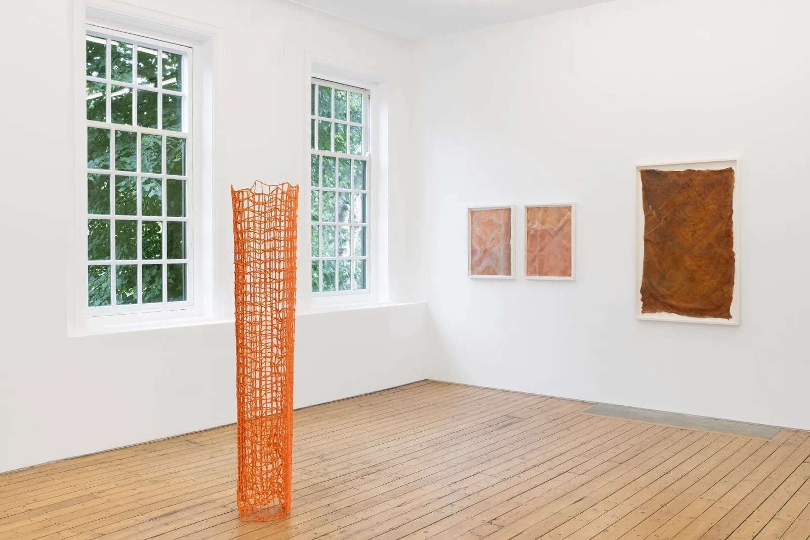 In a white gallery room with two paned windows, three wall pieces made of brown latex hang on the right wall. On the floor space, an orange, netted sculpture arranged in a cylinder hangs from the ceiling and reaches the floor.