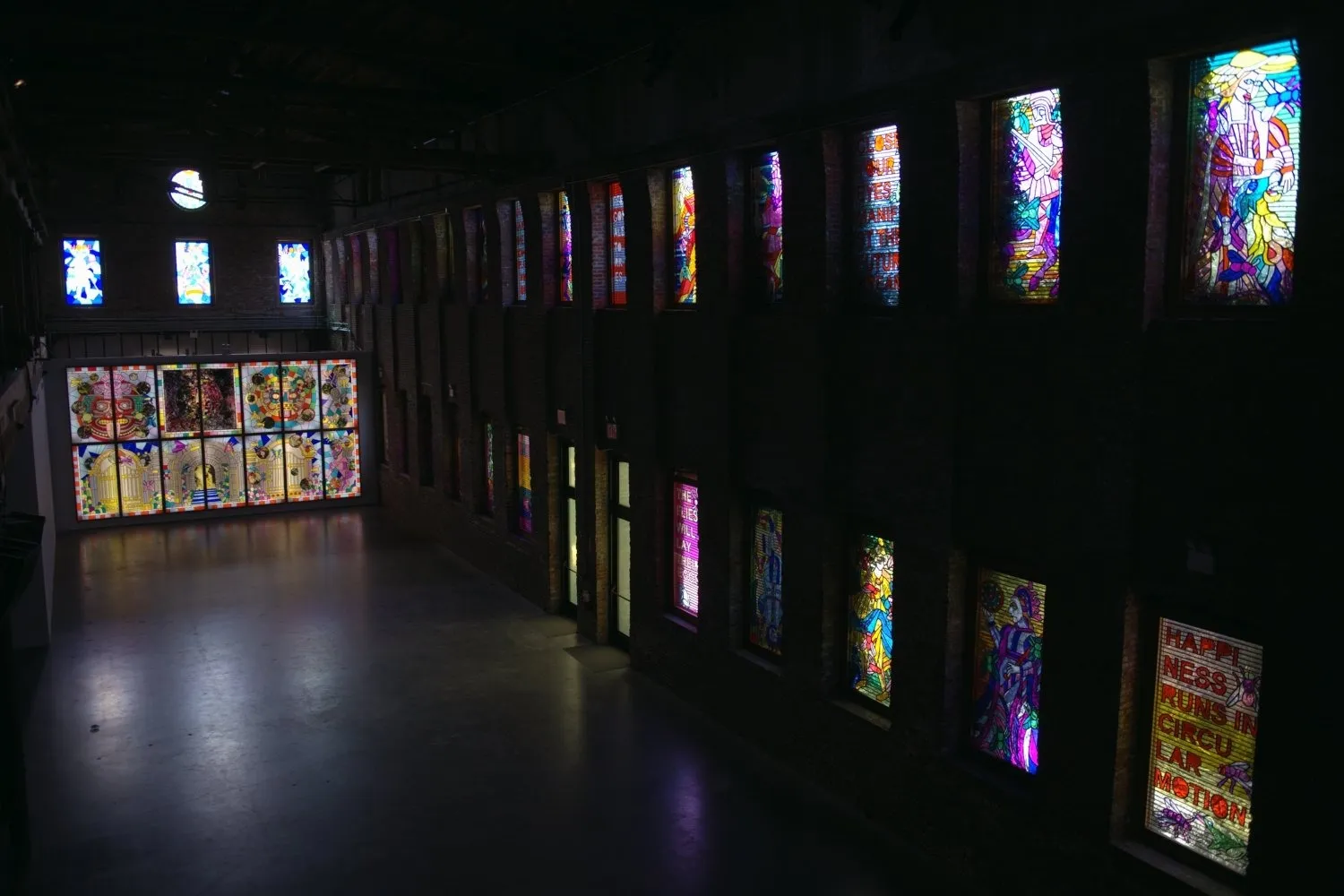 An above shot of a dimly lit building with stained glass in each double-story row of windows. The front features a rose window, three more windows underneath, and a fake wall, backlit and holding a panel of fourteen more windows.