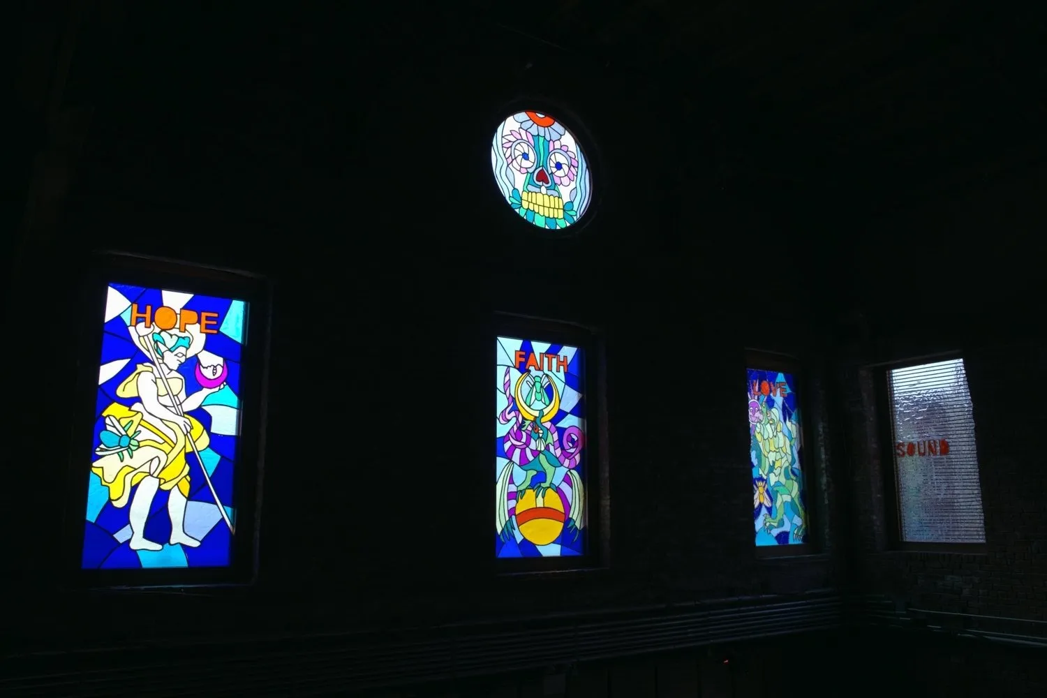 In a dimly lit building, a rose window features a stylized floral skull design in stained glass. Three more stained glass windows below depict different mystical figures with separate words that read "HOPE," "FAITH," AND "LOVE."