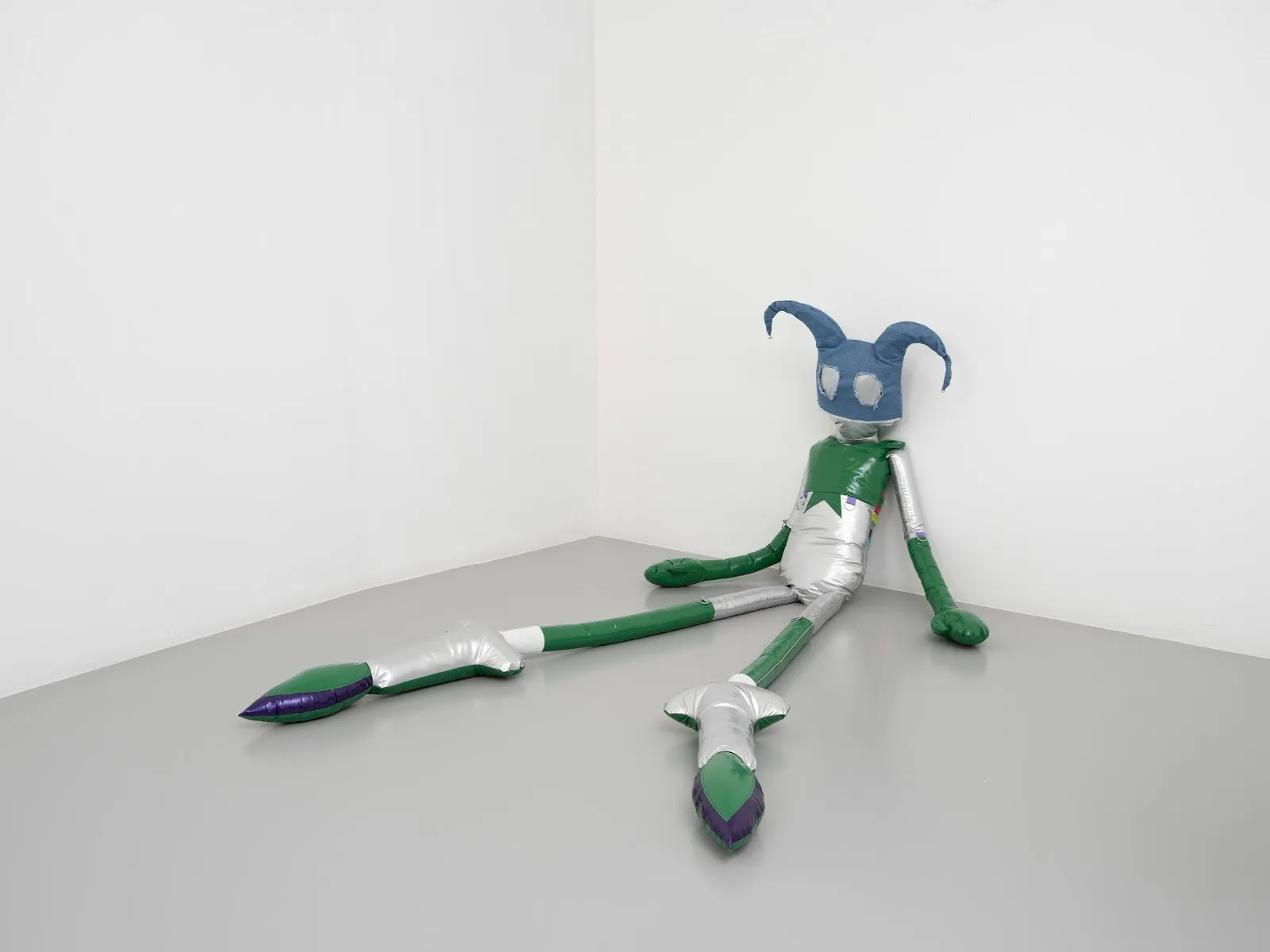 A plastic-coated plushie of sorts, made from sliver, green, purple, and blue tones, slumps against a white wall, its legs extended. The sculpture's head is made in the shape of a blue, harlequin-like headpiece.