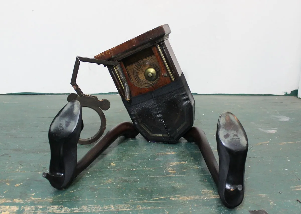 An antique clock tilts to one side, with reused chair legs protruding from the bottom. The legs are adorned with black pumps at the feet. The entire sculpture sits on the floor.