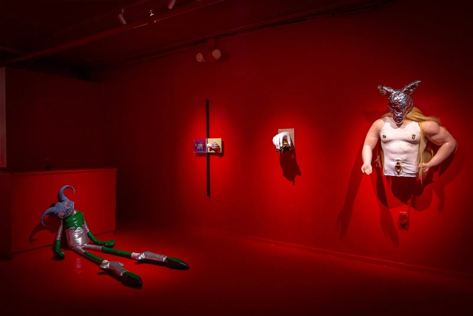 A red-painted room features wall mounted and free standing sculptures. On view is a large, plasticky humanoid plushie, two smaller, 3D wall works, and the torso of a masked figure with visible nipples and a large bellybutton piercing.