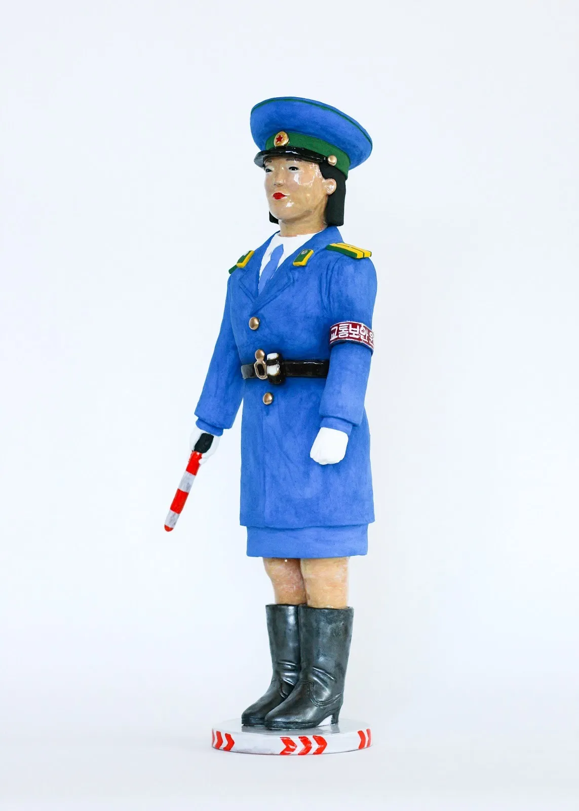 A glossy and matte sculpture depicts a uniformed guard in blue, standing straight and holding a crossing stick/sign.