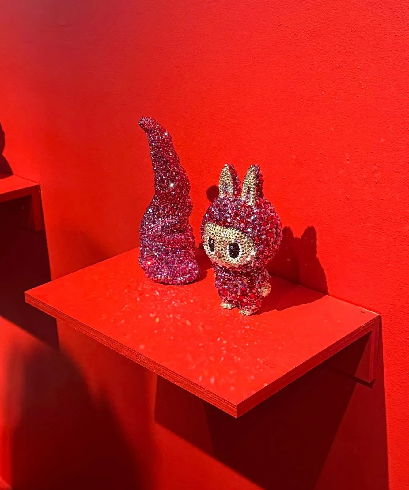 On a small red shelf mounted to a red wall, two pink bedazzled objects sit. The left is a tentacle of sorts, sitting upright. The right is a jewel-encrusted Labubu.