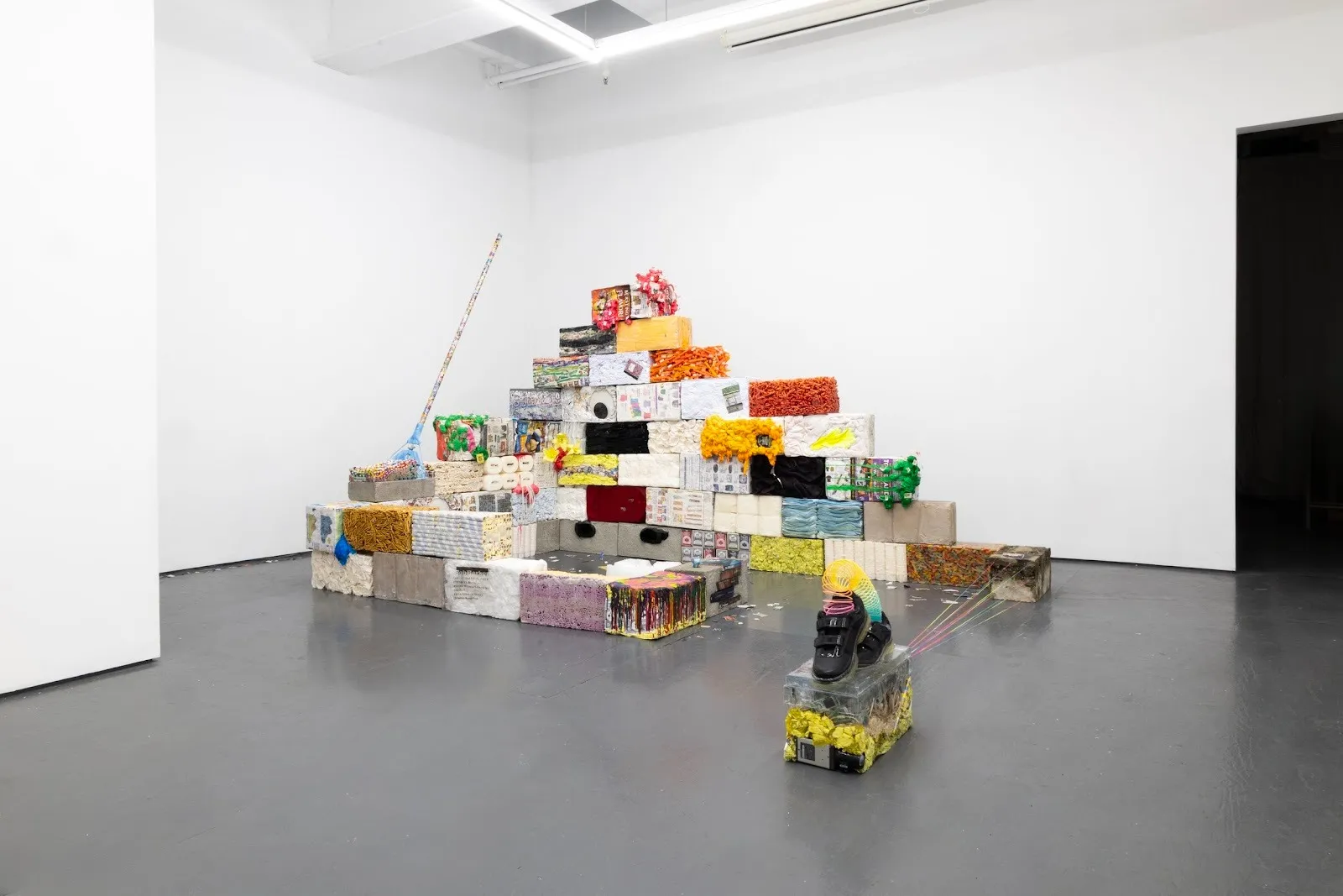 In the corner of a white gallery room, various colorful blocks made of different material are stacked on top of each other and arranged in a square.