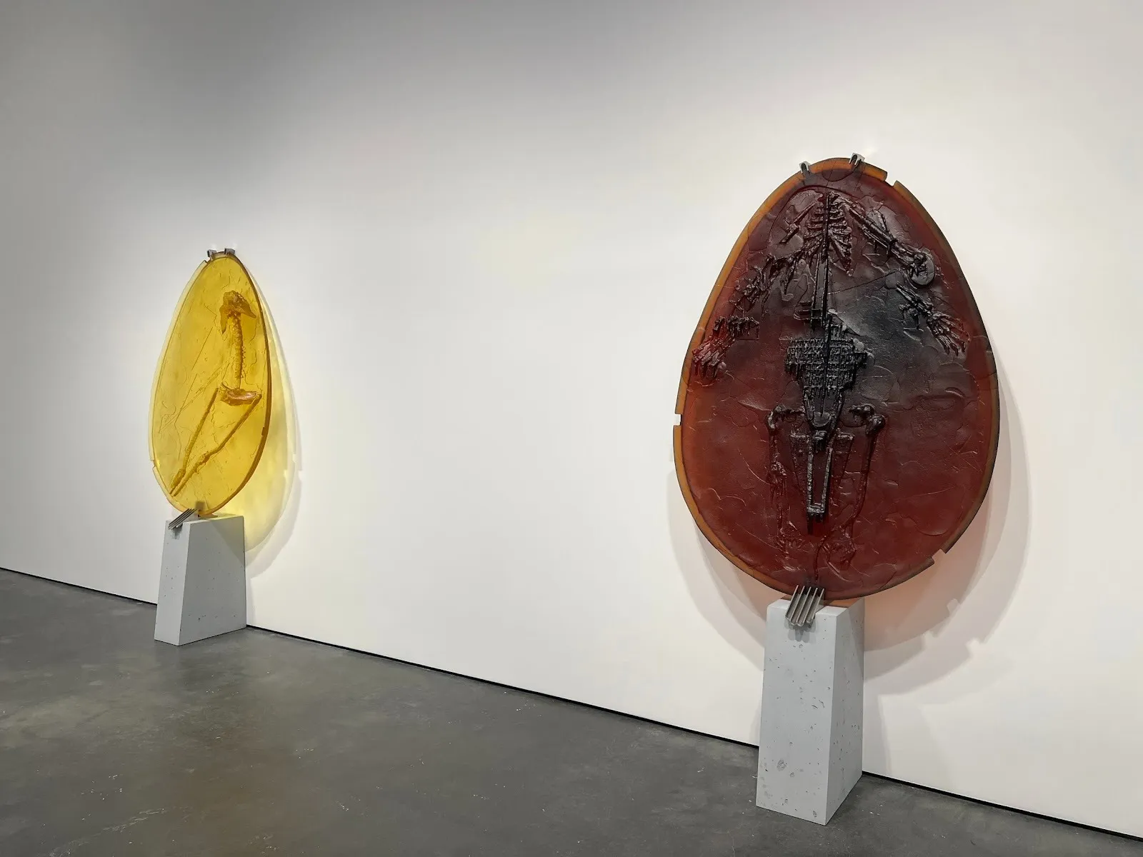 Two sculptures placed on small pedestals flush to the wall lean against a white gallery wall. The sculptures appear as thin, glassy egg-shaped slabs, with designs trapped inside and protruding outwards, texturing the surface.