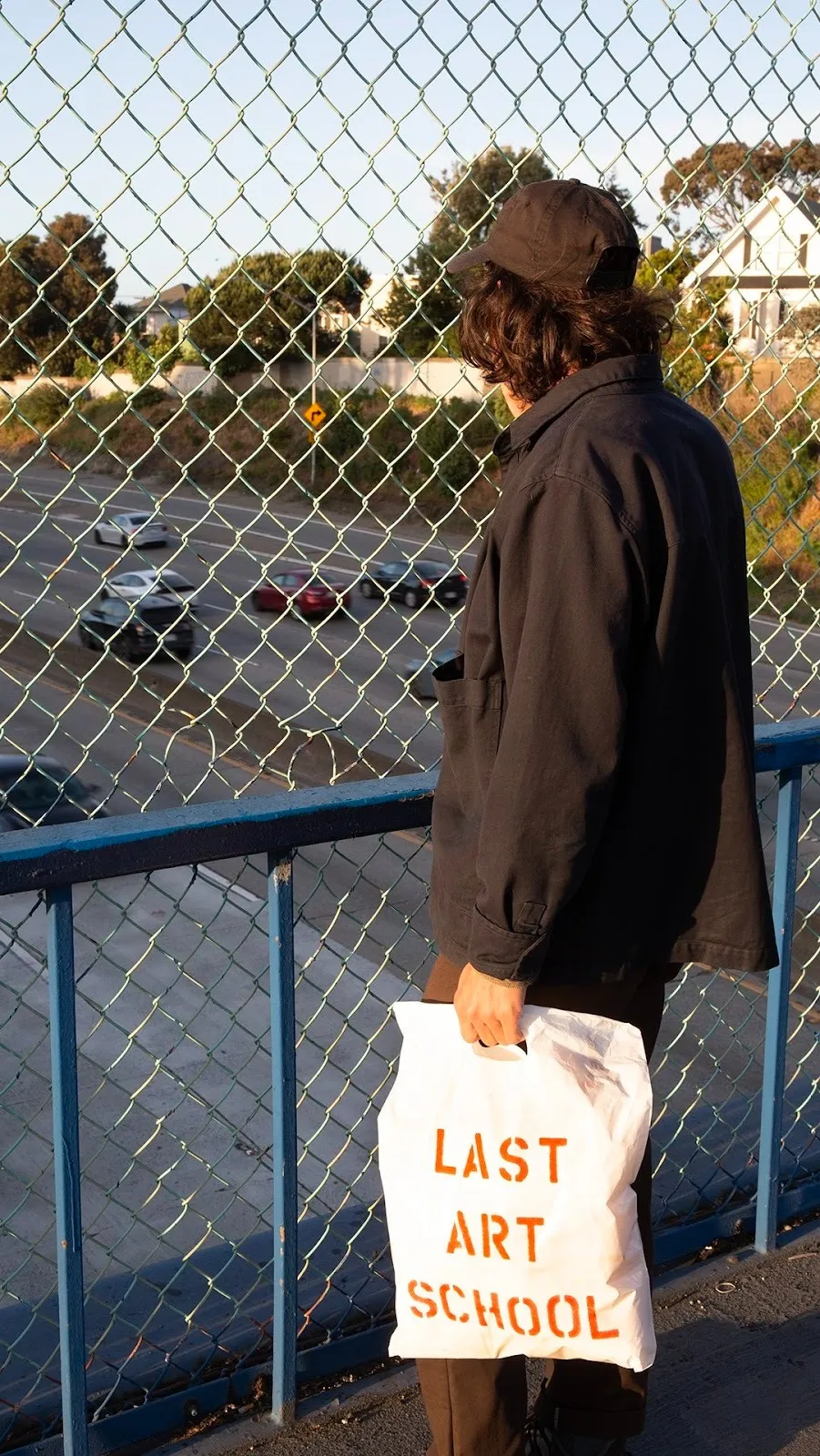 On a freeway overpass, a person clad in black faces and looks past the fence and carries a white bag that reads "LAST ART SCHOOL" in red text.
