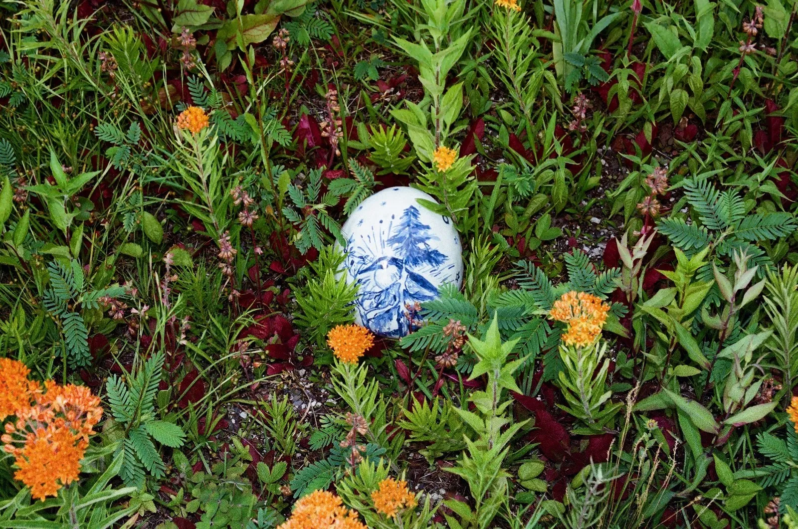 An ornately decorated blue and white egg nestles in small, orange flowers and leafy grass.