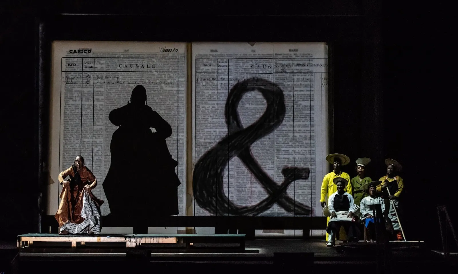 A stage features a backdrop of paper pages with small, crowded text. The right page is covered by a large ampersand. One performer to the left sings and motions, their silhouette projected on the backdrop. An chorus-like ensemble gathers stage left.