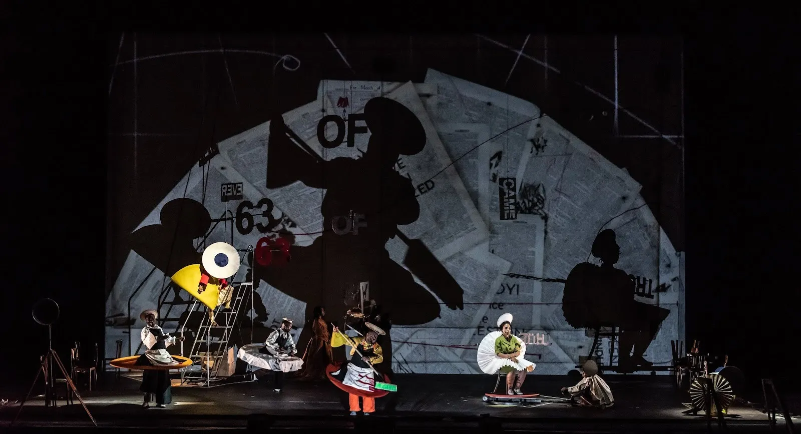 A stage features a backdrop of scattered leaves of pages. Multiple performers engage in a variety of motions: sitting, singing, handling props, dressed in colorful, voluminous costumes.