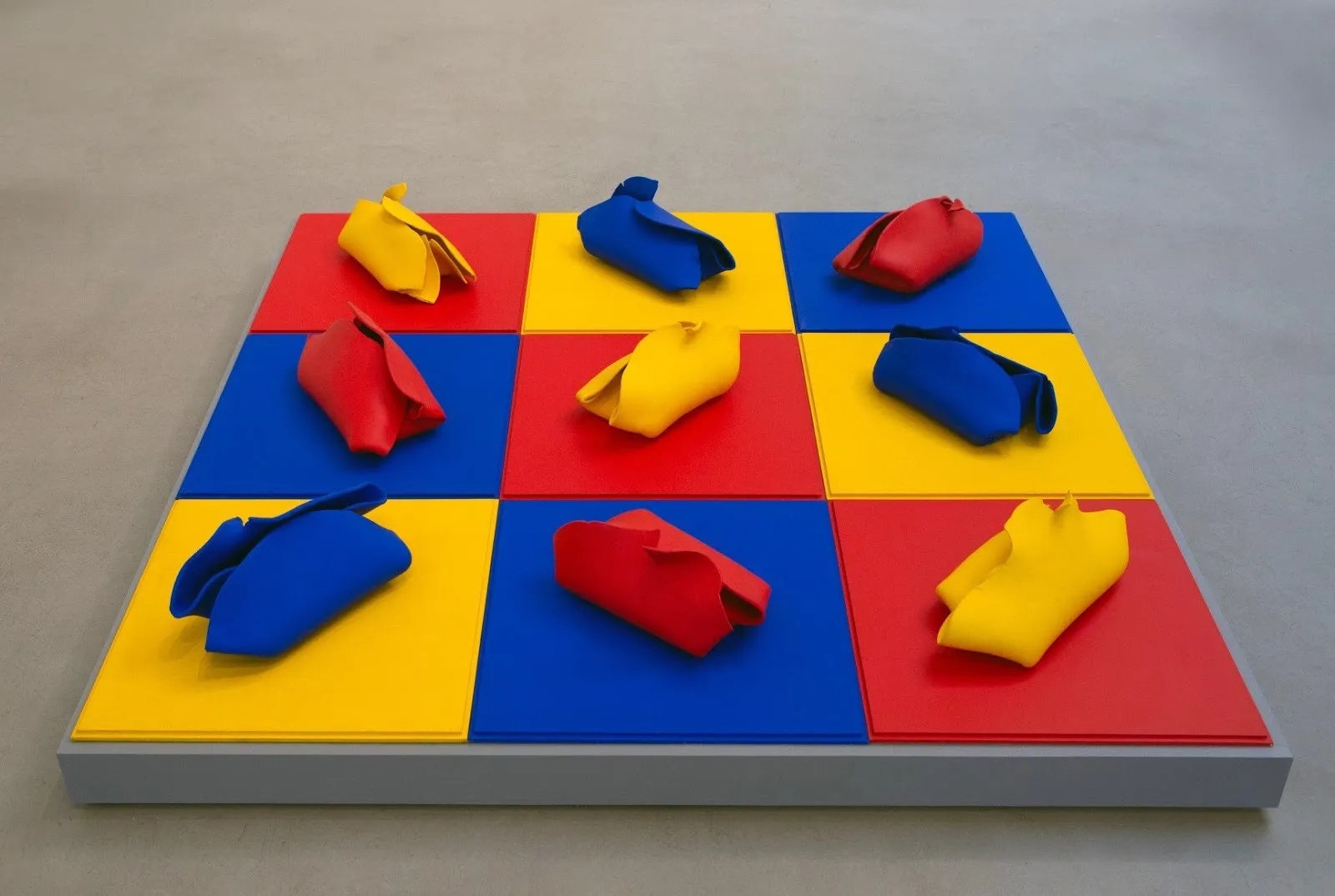 A sculpture is made from a raised square platform and features a checkerboard grid of red, yellow, and blue squares. On top, Red, yellow, and blue folded sculptures rest on each square, mismatching in color with the square underneath.