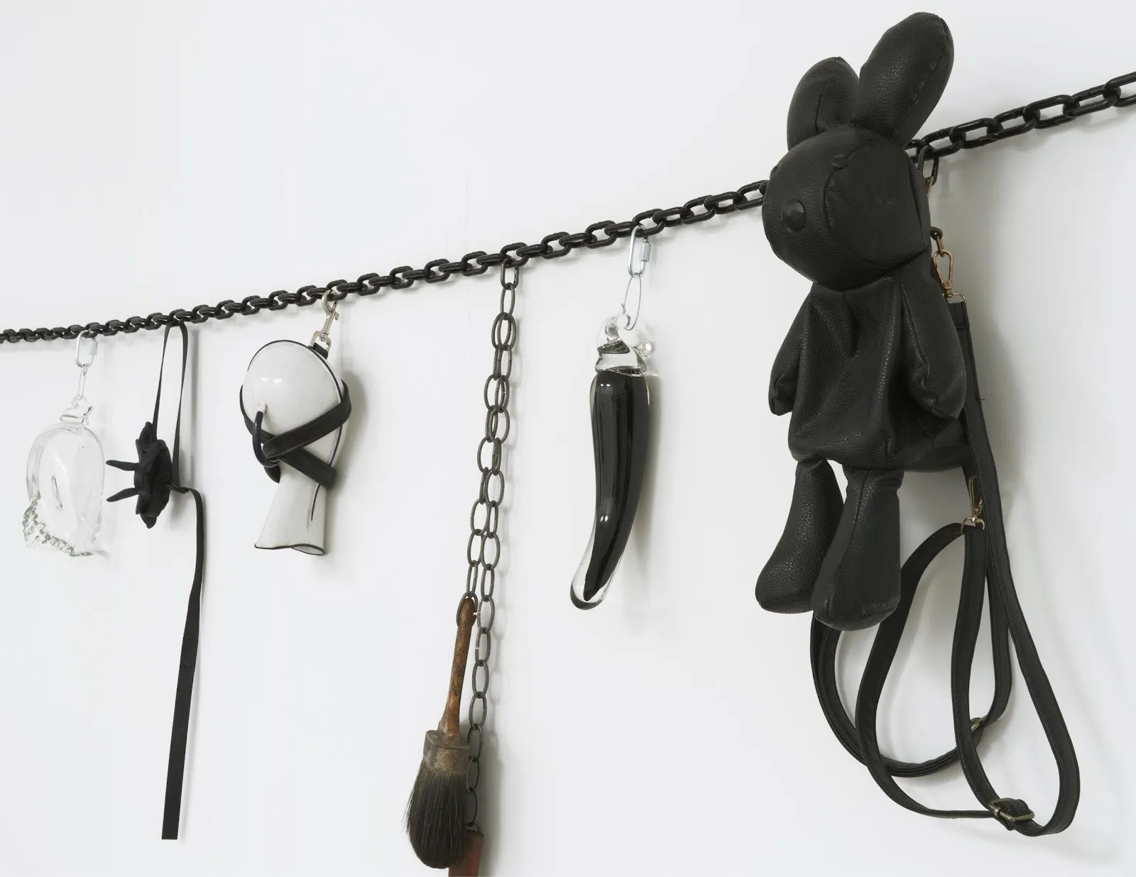 A black chain strung against a white wall supports a variety of clipped-on attachments. The attachment objects include a black leather bunny, black straps, and a glass phallus.