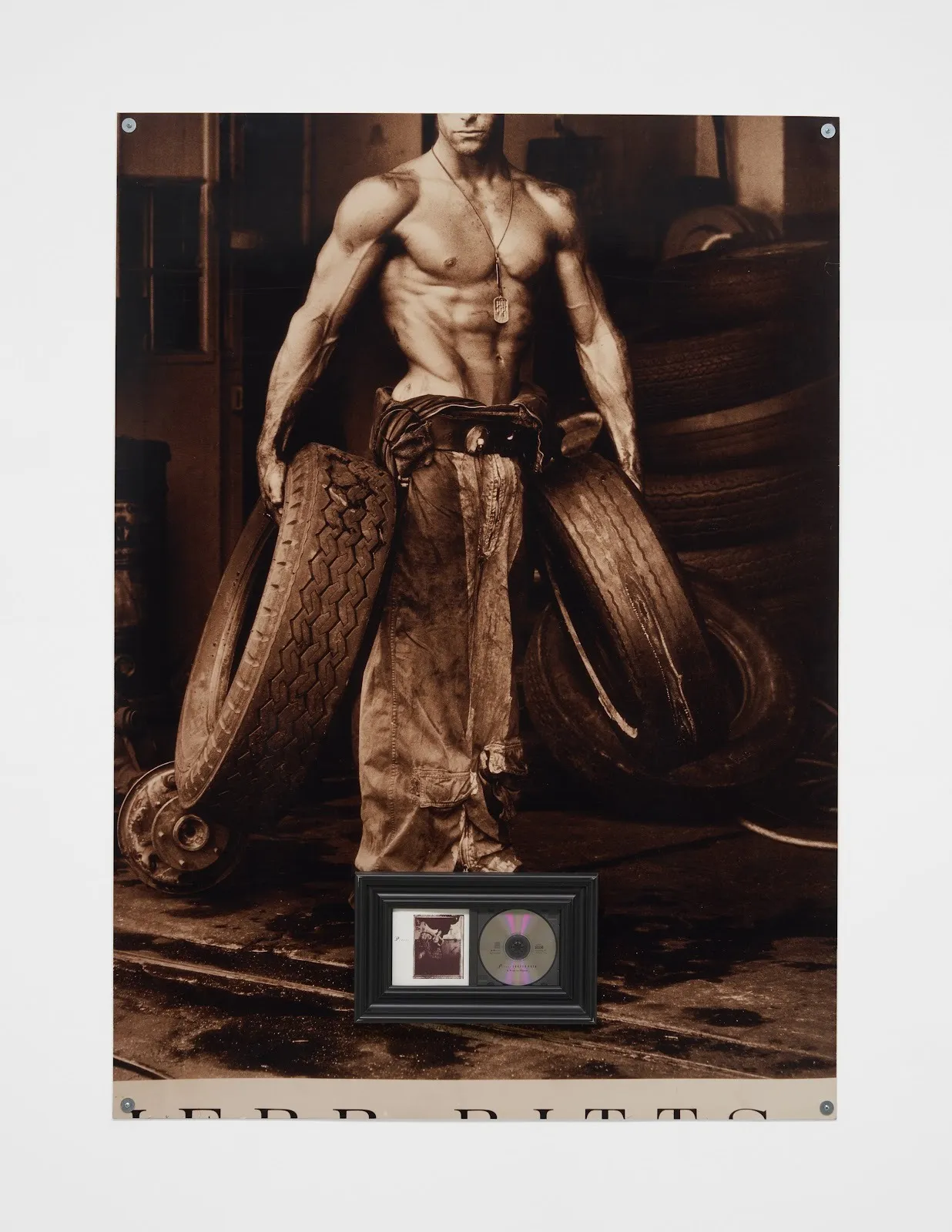A 2D work pinned to a white wall features a sepia photograph of a muscled man carrying a tire in each hand. Towards the bottom, an embedded black frame contains a small sepia photograph and a CD.