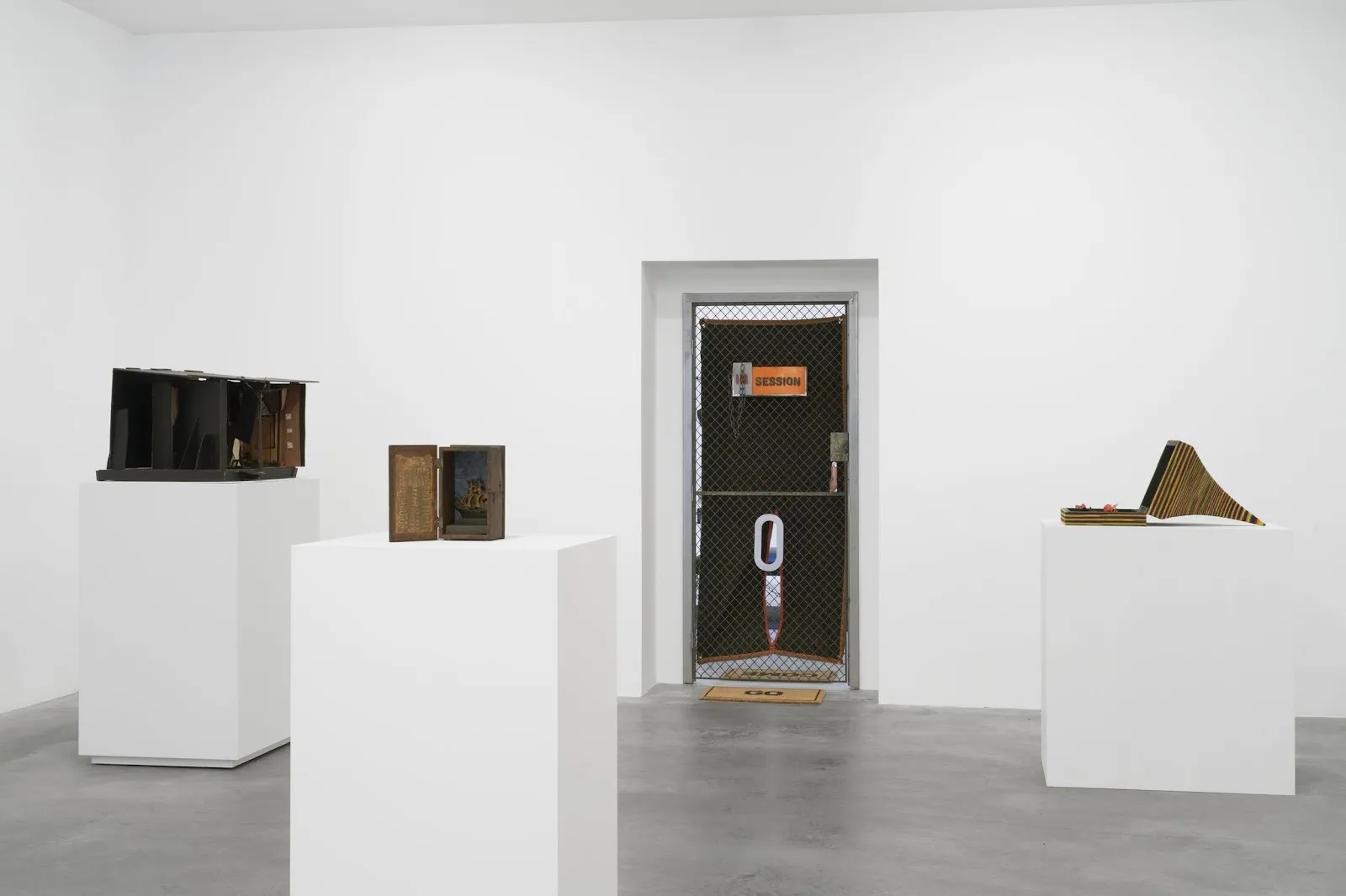 In a white gallery room, three white square plinths support boxy sculptures. The gallery's back door is made of chain mail, black tarp, a push bar, and a sign that reads "SESSION."