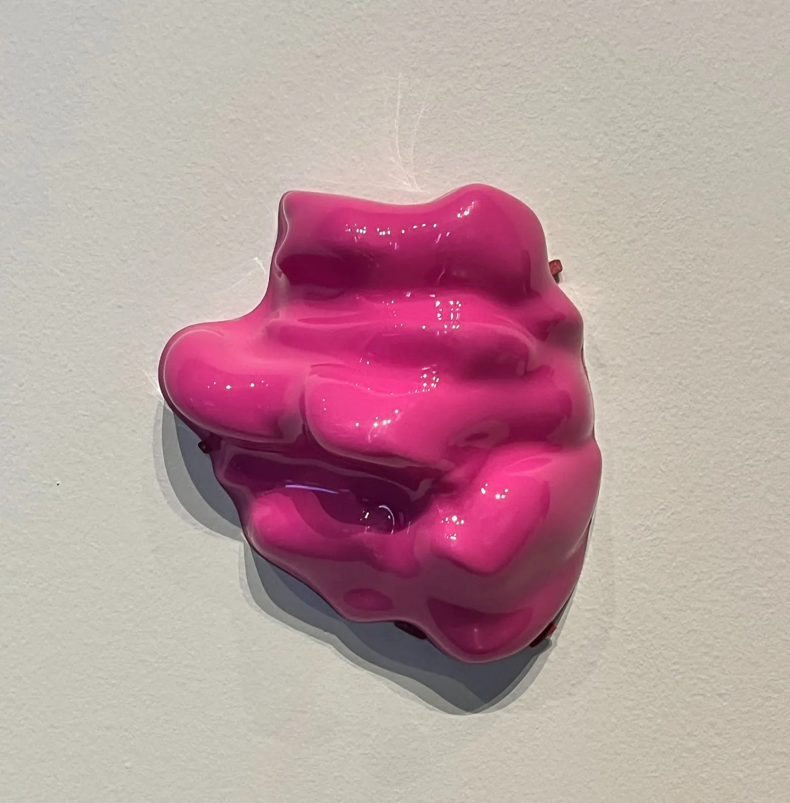 Mounted to a white wall, a shiny, plasticky sculpture is molded into a bright pink, lumpy glob shape.