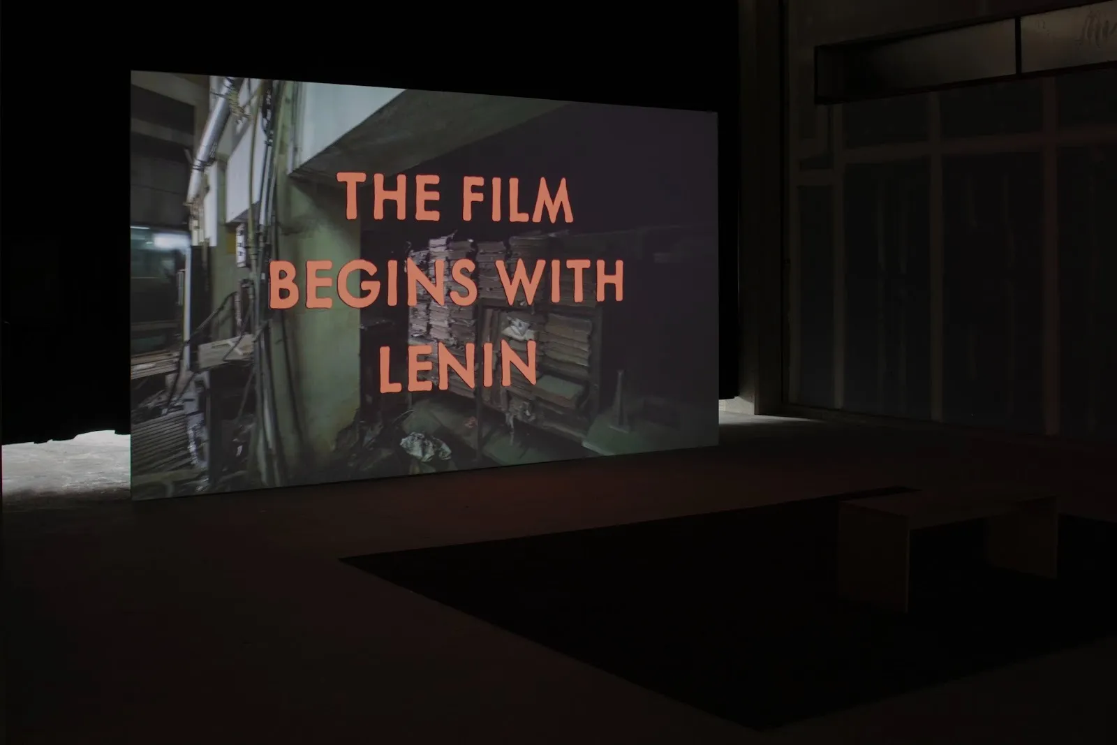 A dark, curtained room hosts a large projection screen. The screen features an industrial, office-like space overlaid with large words in orange that read "THE FILM BEGINS WITH LENIN."