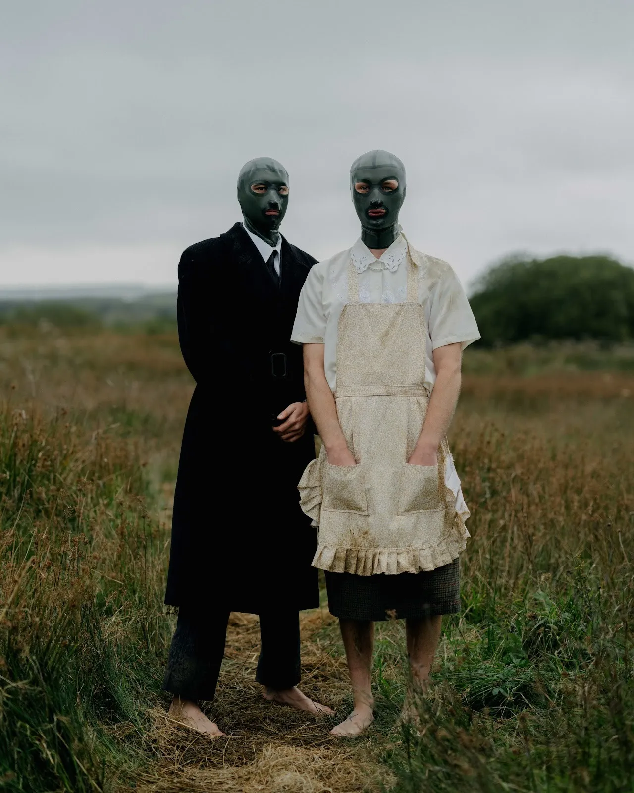 Two barefoot figures, one in a black trench coat and one in a white blouse and apron, stand in a grassy field on a cloudy day. Both figures wear dark latex balaclavas that have two eye hols and a mouth opening. The figures look directly at the camera