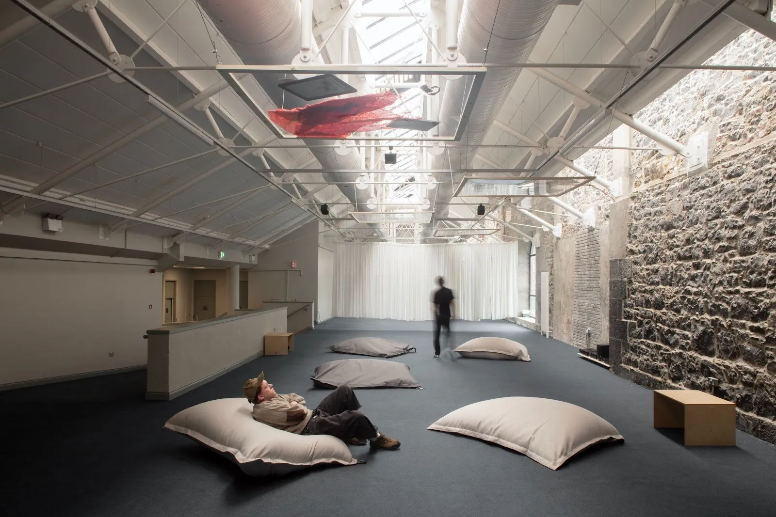 A large room with white piping and beaming and a skylight is filled with four large pillows on the ground. A person reclines, looking up at a glass frame filled with media, hung horizontally from supporting beams.