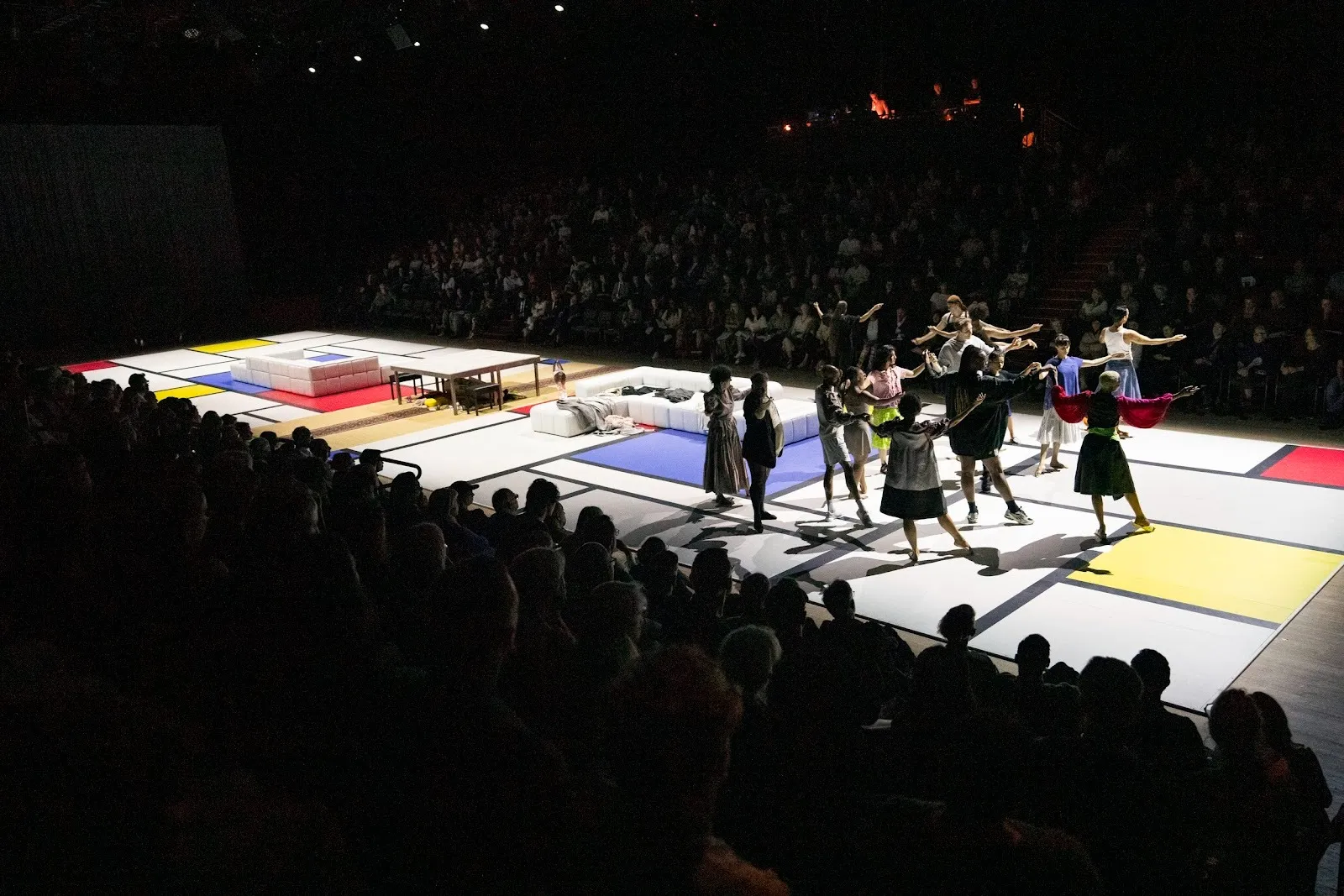 On a spotlit Mondrian-esque runway staged with white couches and a table, a group of performers gather and perform a choreography, their arms extended. A large audience lines each side of the runway.