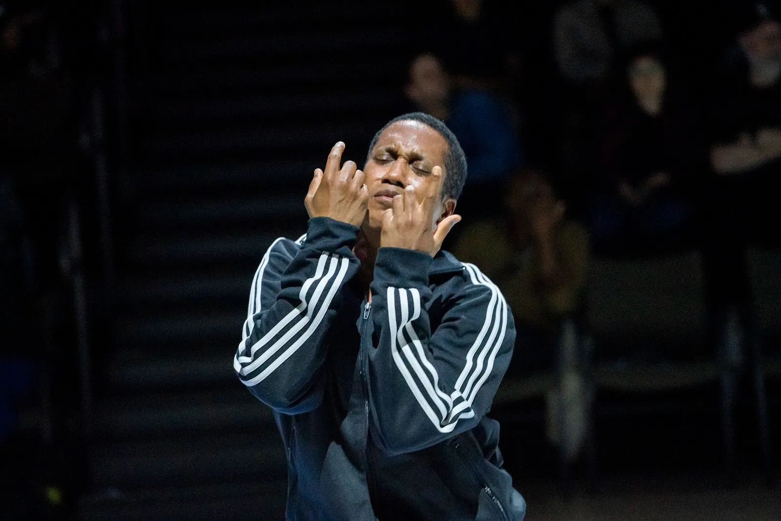 Against a background of a dimly lit audience, a man in a black adidas tracksuit performs a choreography, eyes closed, hand raised to his face, body slightly curved and contorted.