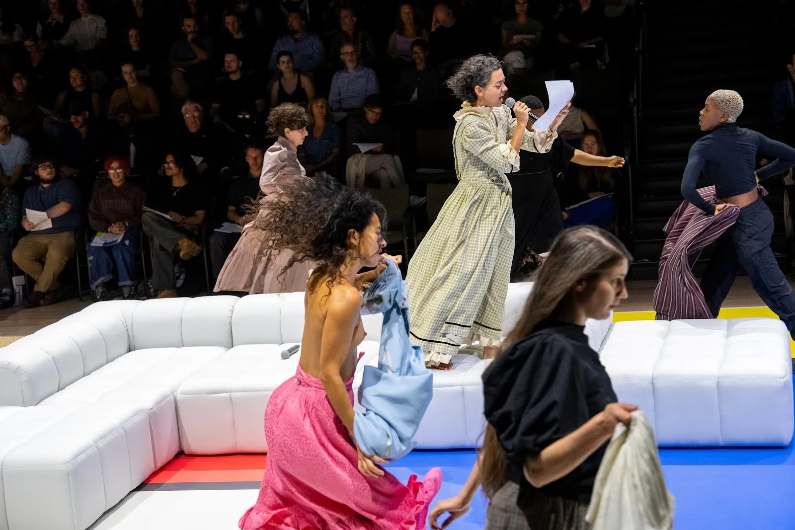 Performers on a yellow, white, red, and blue runway, staged with a white couch, move energetically about, each undressing or holding clothes. One performer stands on the couch and reads from a piece of paper. An audience watches from the background.