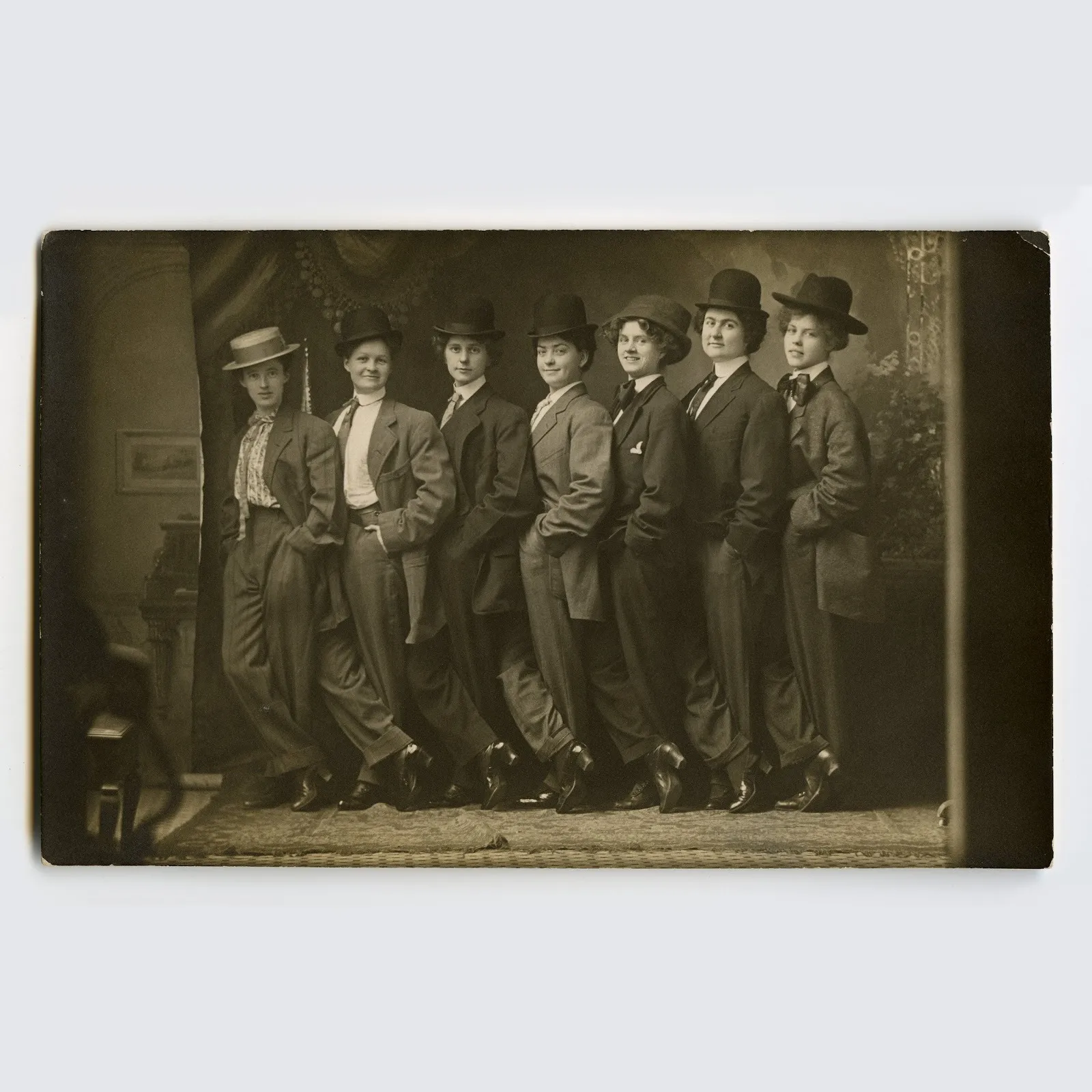 A vintage black and white photograph captures seven women, posed and lined up with hands in their pockets, wearing men's suits and hats. The photograph appears to be taken in a photography studio.