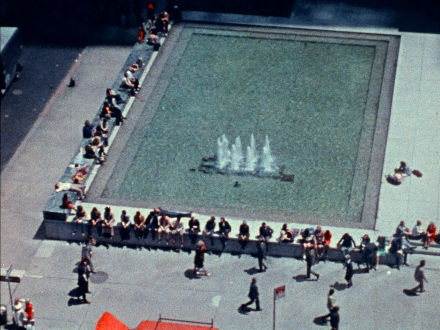 In an aerial shot, many people sit on the edges of a large, rectangular fountain set in a large city square/courtyard.