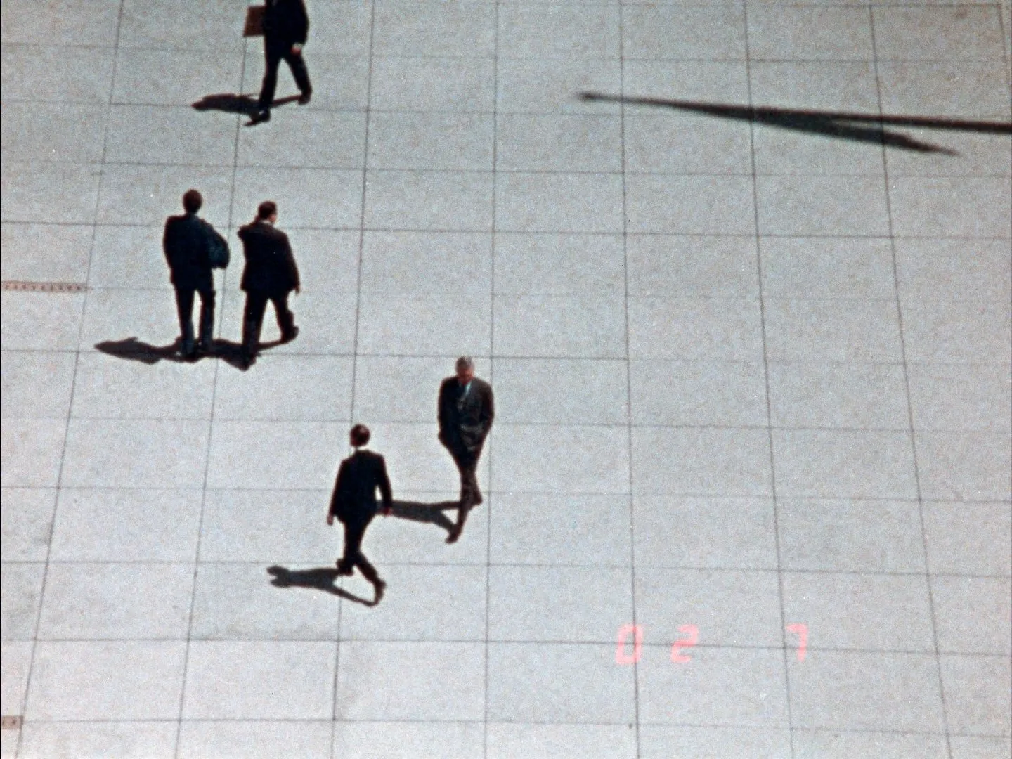 Four men in business suits walk disparately across a wide, stone-tiled square, casting long shadows.