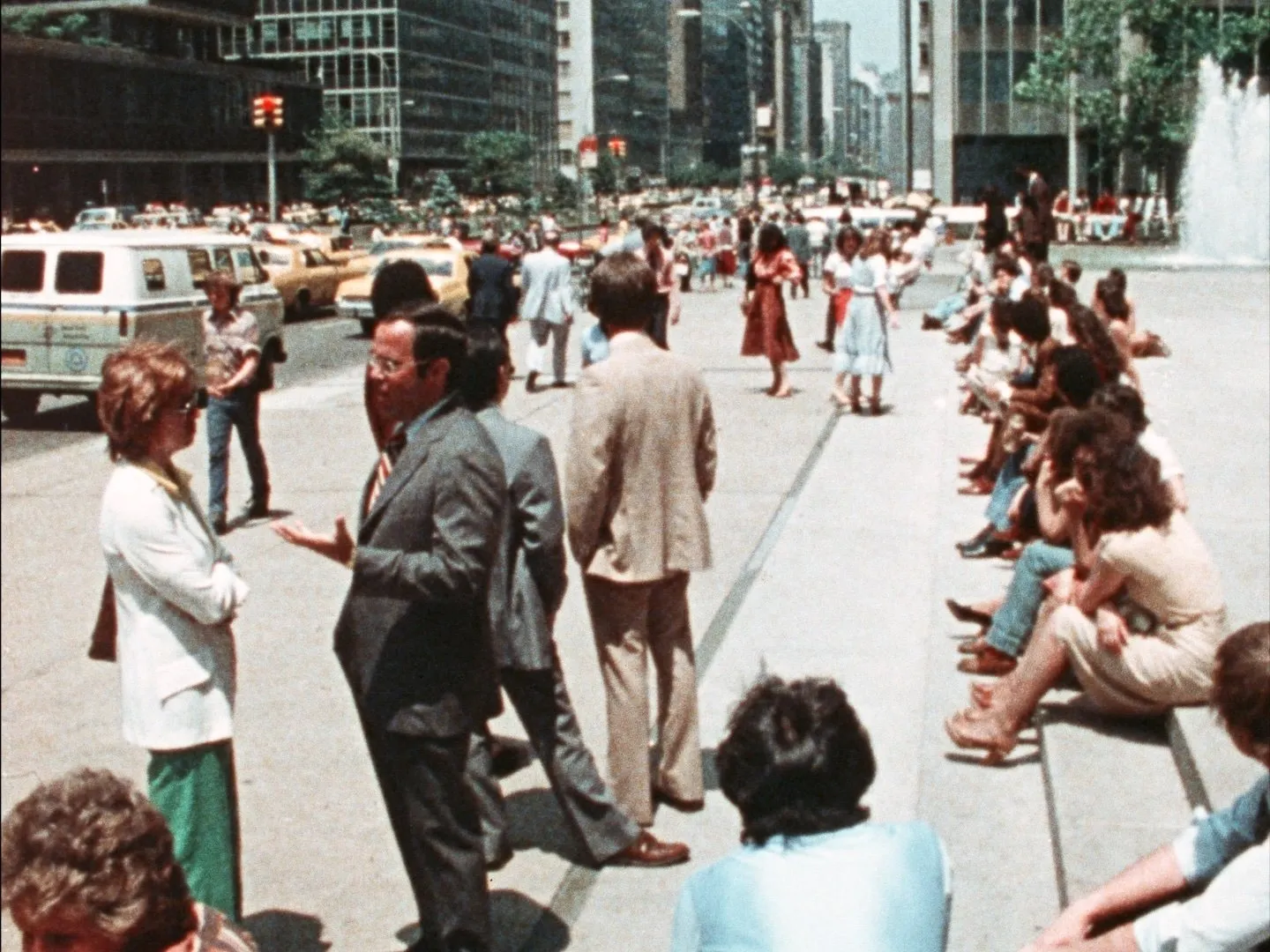 On a busy city street, cars drive along the asphalt, while crowds of people sit and stand on the adjoining sidewalk.
