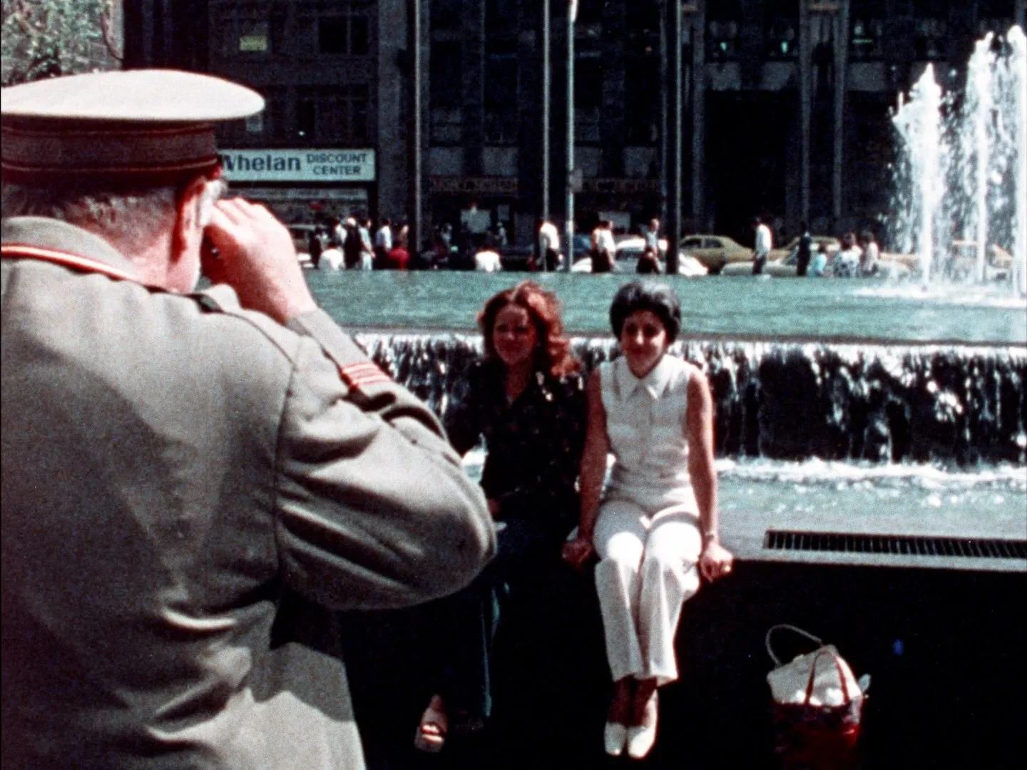 With his back facing to the camera, a man takes a picture of two women sitting on the edge of a large, public fountain in a busy city.