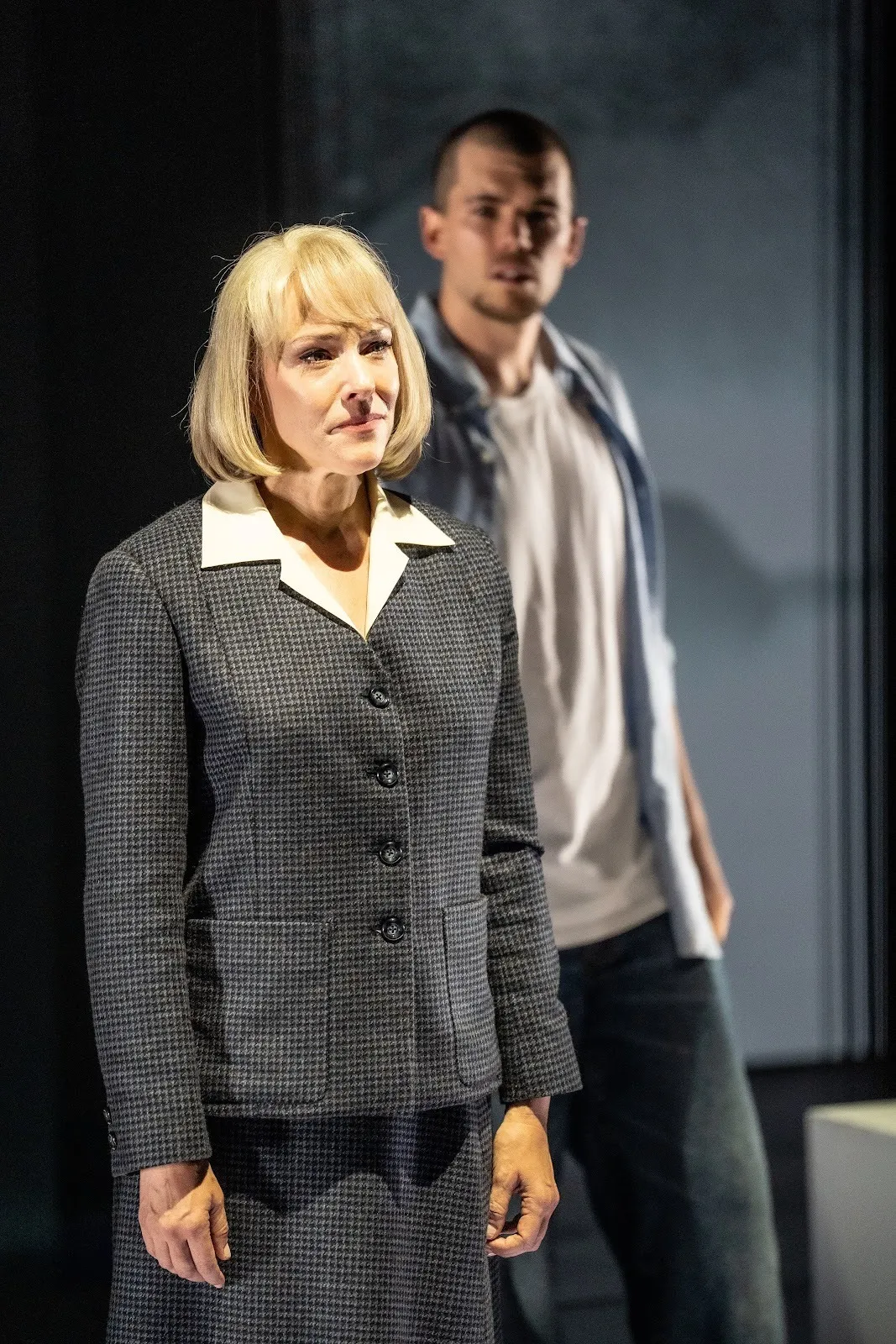 A woman in a tweed suit with a blonde bob looks to the side off-camera, while a man in a blue polo button down looks toward her in the background.