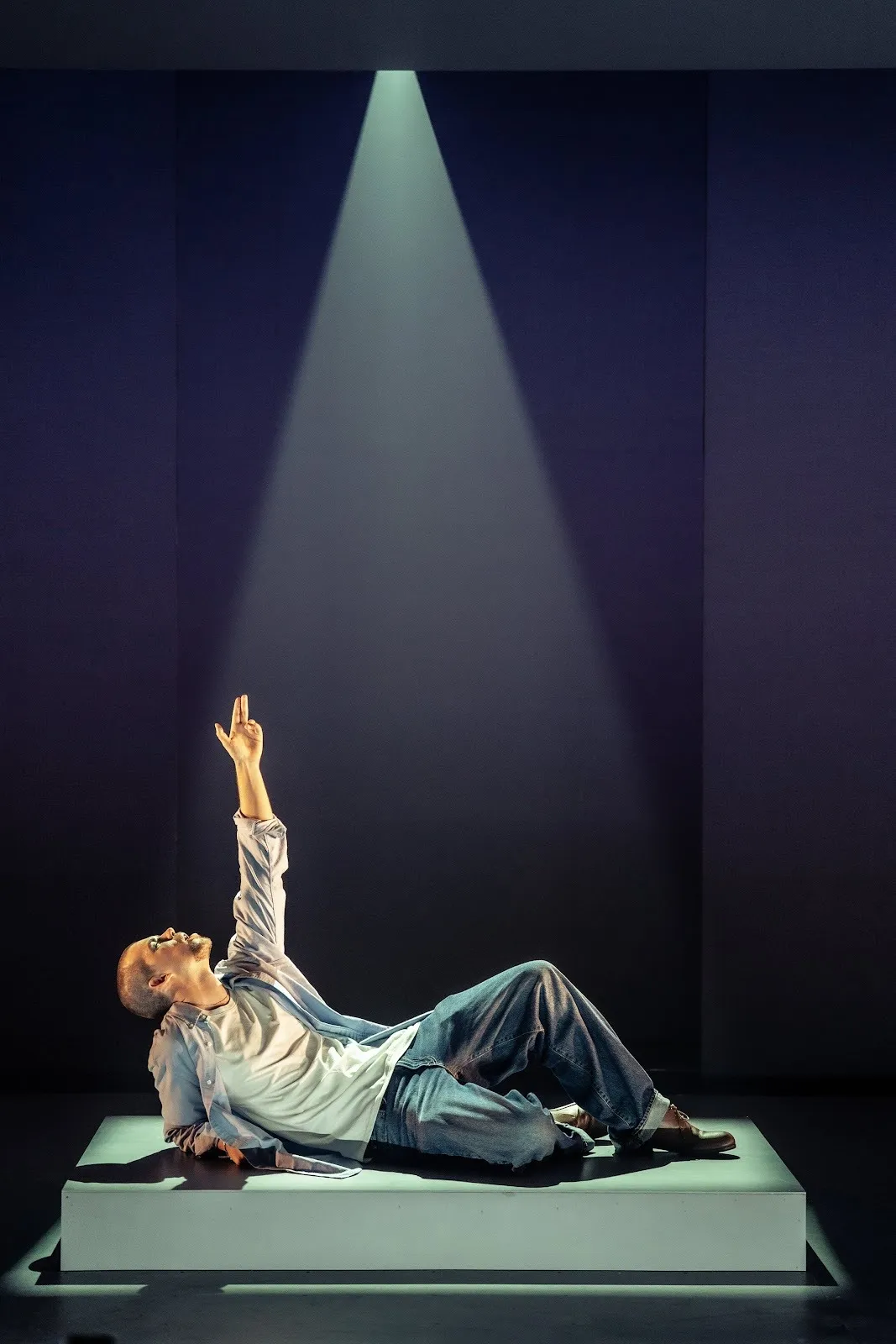 A man in a blue polo shirt lies on a platform and props himself up with one hand, the other raised to the ceiling. His face also looks to the ceiling. A single spotlight illuminates him.