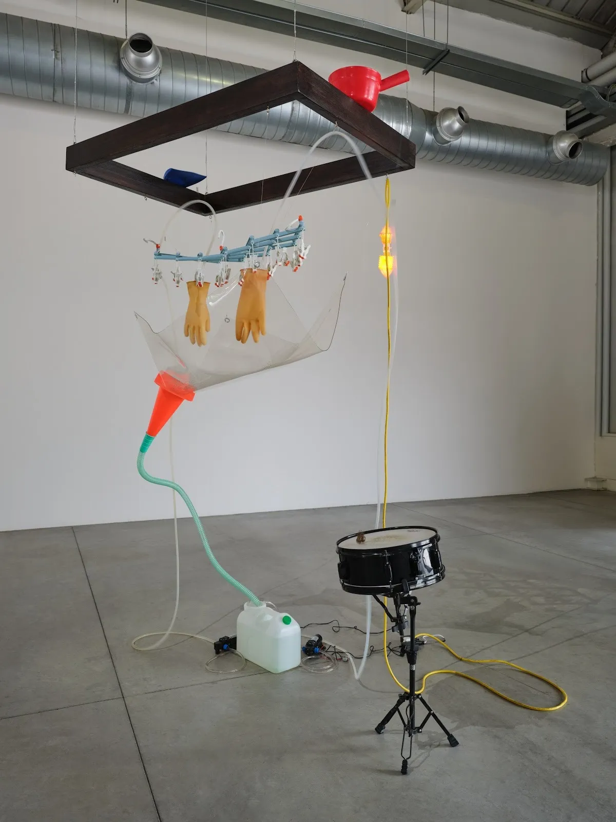 A wooden frame hangs from the ceiling and supports a large red cup/ladle. Attached and hanging is a plastic, tubing, glove, cone apparatus hooked to a lightbulb, jug, and snare drum.