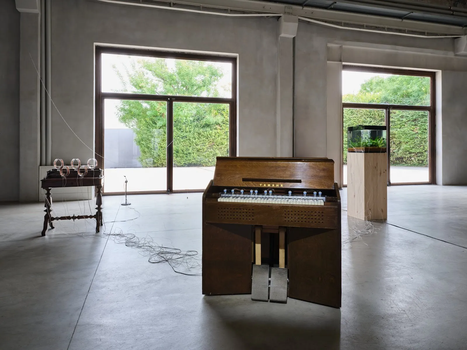 In a concrete warehouse with large windows, a small, antique table, wall piano, and fish tank are all placed away from each other but linked by a variety of wires.