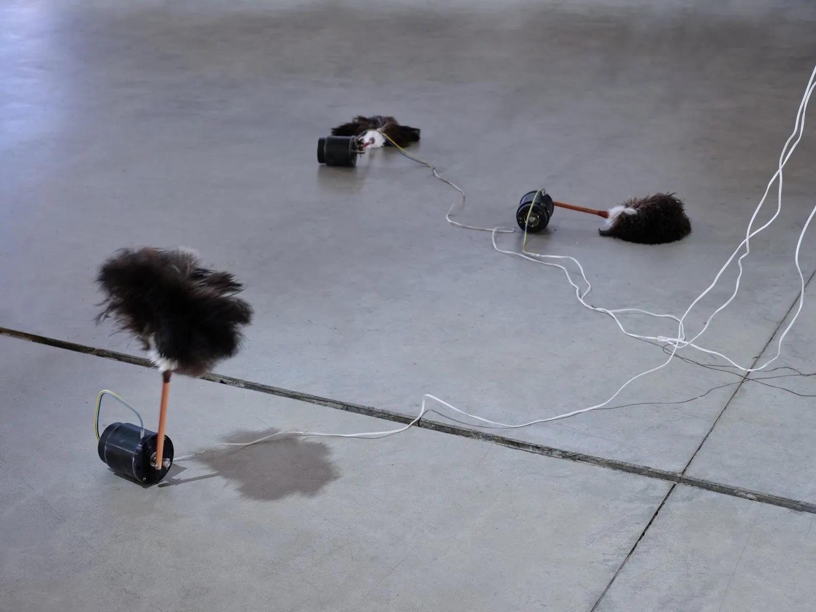 On a concrete floor, feather dusters attached to motors and wires move or lie on the floor.