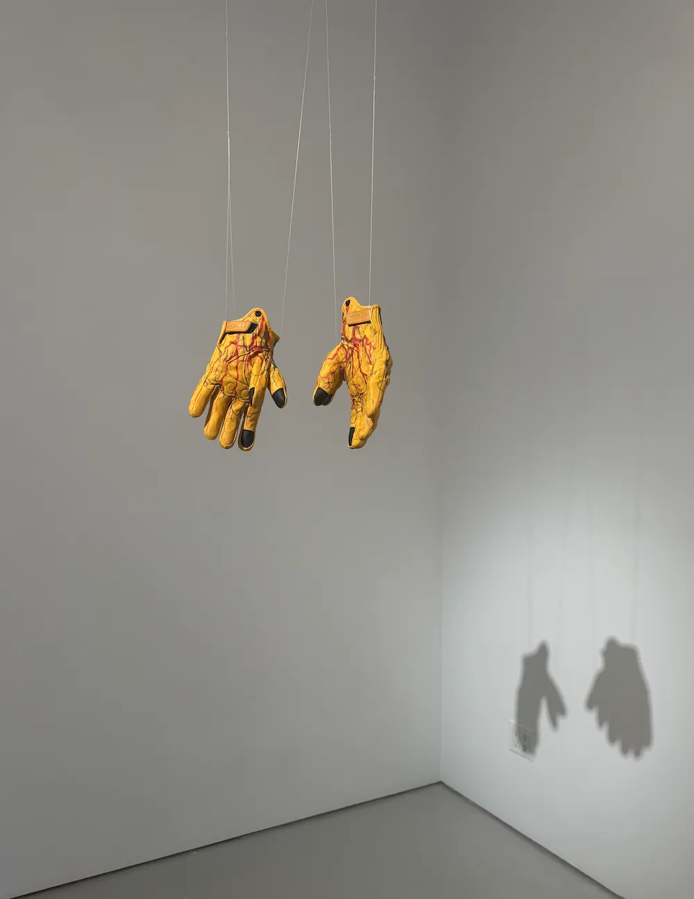 Two yellow work gloves with painted red veins suspended from the ceiling on thin wires.