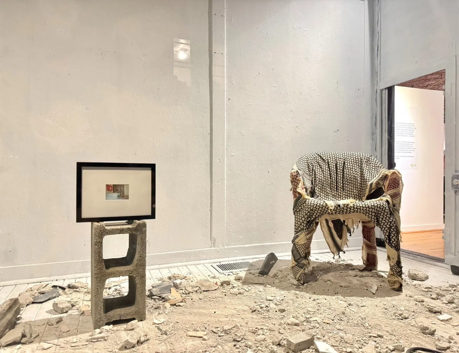 Installation with a chair draped in patterned fabric and a small framed image on a cinder block, surrounded by rubble on the floor.