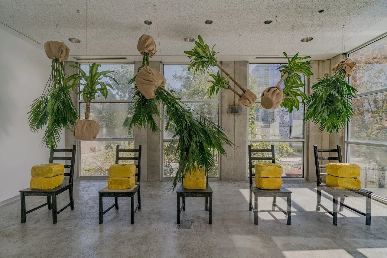 Installation with black chairs topped with yellow blocks and suspended plants wrapped in burlap, hanging upside down in a bright gallery space.