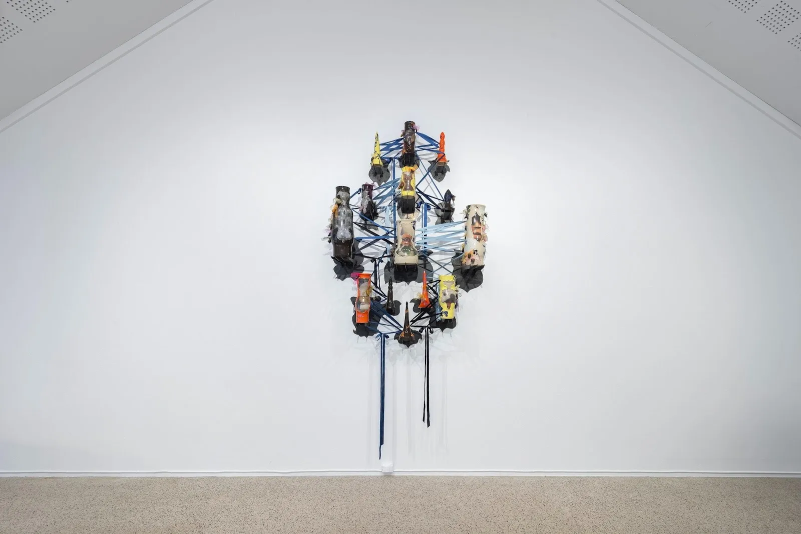 Mounted to a white wall, various ceramic structures are threaded by tangles of blue ribbon.