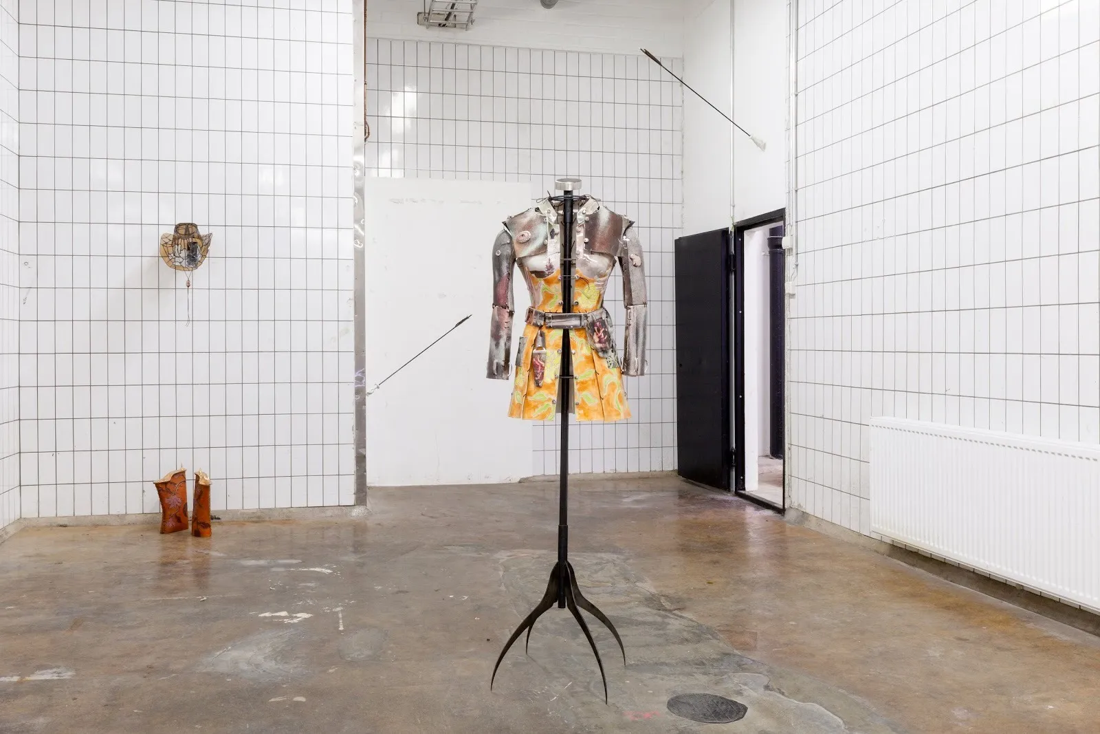 In a tiled white room with shiny concrete floors, various works stand on the floor or are mounted to the walls. The central piece is a mannequin dressed in both a decorated orange bodice and skirt as well as shoulder and arm armor.