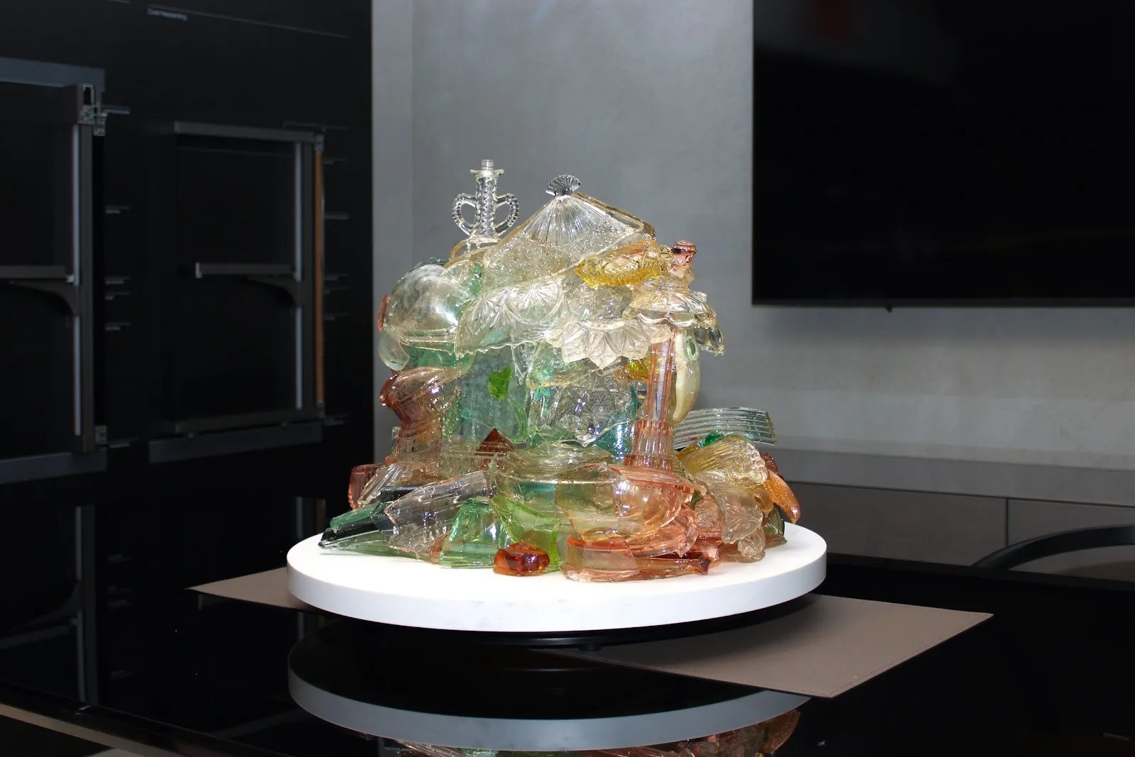 A glass sculpture appears as an amalgamation of multiple disparate pieces and forms: some from clear, red, blue, red, and green glass. Some recognizable shapes appear as bottles and decoration. The pieces pile together on a white circular plinth.
